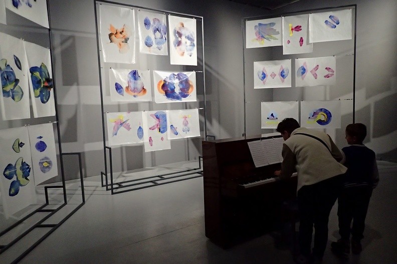 44-P9140093 -- Sala Muzyczna.
Na stelażach wisi "partytura, oparta na cyklu obrazów <i>500 Azaleas</i> namalowanych przez Alexa Cecchettiego. [...]
Partytura utworu jest gotowa, więc każdy z odwiedzających może go wykonać na pianinie, by posłuchać,
jak brzmią kolorowe rysunki artysty przetransponowane na zapis nutowy"