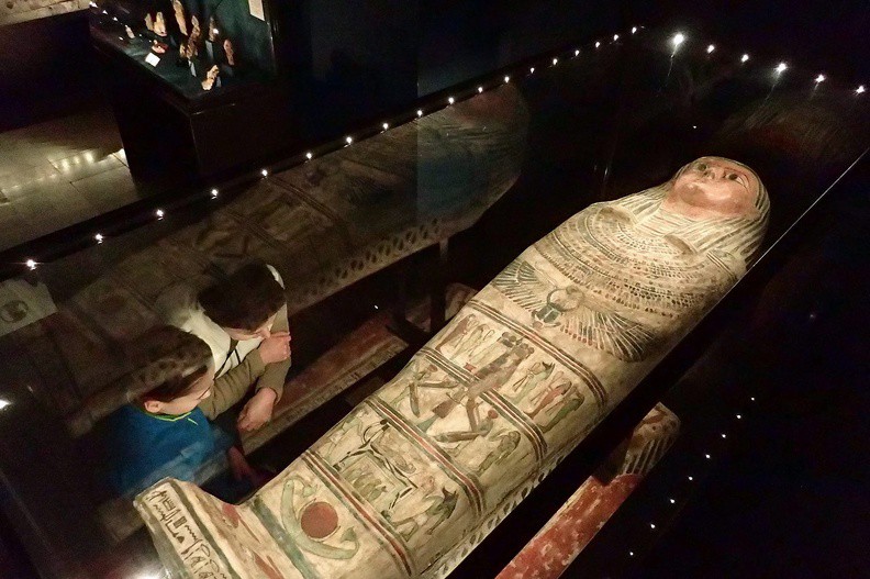 045-P1080264 -- Sarkofag z el-Hibeh, pochodzący z późnego okresu ptolemejskiego bądź wczesnorzymskiego (lata 332 p.n.e.-395 n.e.). W trzecim od dołu rejestrze (ilustrowanym "pasie") znajduje się symbol boga Ozyrysa, po którego bokach stoją boginie Izyda i Neftyda. Widoczna z prawej strony kobieta uniosła dłoń do ust – jej gest oznacza żałobę. Rejestr przedostatni wyobraża łoże balsamierskie, na którym spoczywa zmumifikowany Ozyrys; bóstwa ze zwierzęcymi głowami to Anubis oraz Horus. Poziom najniższy zawiera słońce z półksiężycem;
na pokładzie papirusowej łodzi dryfują przez praocean Nun. Zdjęcie w większej rozdzielczości: <a href="http://www.dziennikgorski.type.pl/zdjecia/pojedyncze/P1080264.jpg" target="_blank">LINK</a>